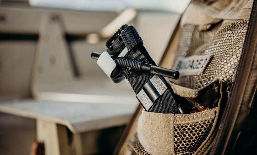 En taktisk ryggsäck med ett fack märkt 'BANDAGE' som innehåller en svart tourniquet från SAM Medical, fotograferad i närbild i en militär eller fältmedicinsk miljö.
