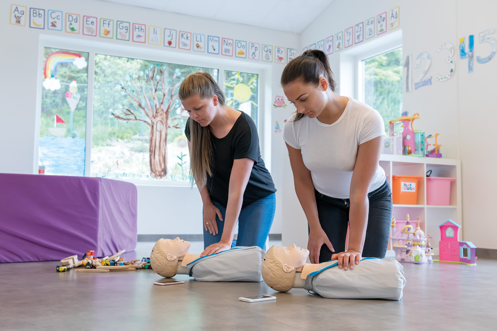 Två kvinnor som arbetar på en förskola övar HLR på varsin Little Junior QCPR HLR-docka, omgivna av leksaker, affischer och andra detaljer typiska för en förskolemiljö.