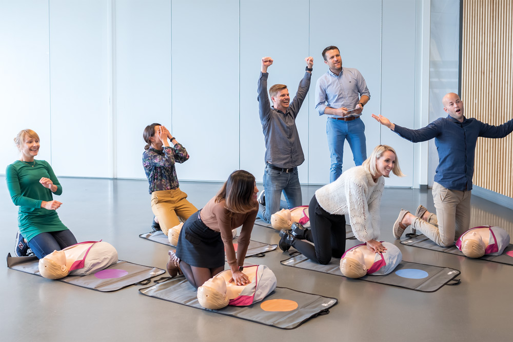 En grupp kollegor övar HLR på träningsmattor med varsin HLR-docka av modellen Little Anne QCPR. De ser både glada och trötta ut efter en tävlingsinspirerad övning.