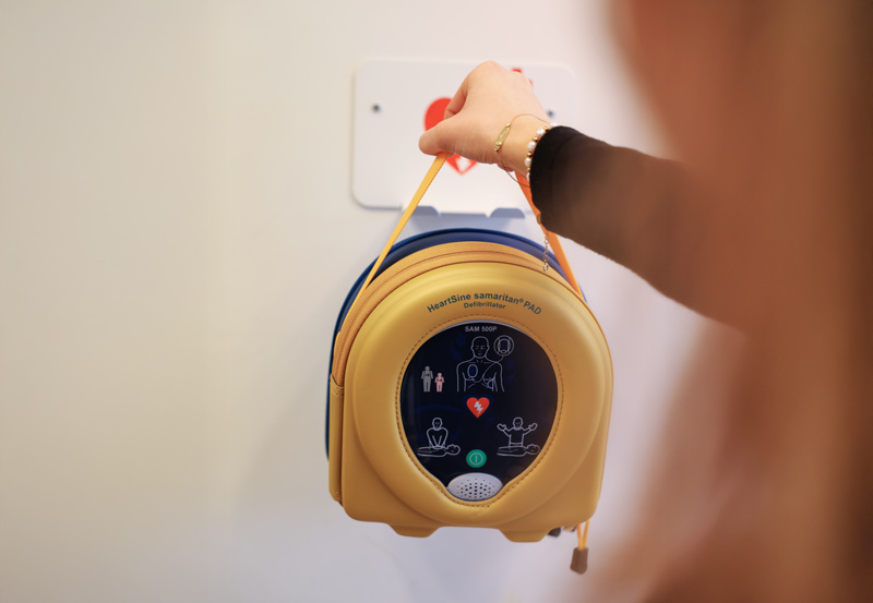 Kvinna i svart tröja hänger upp en hjärtstartare av modellen Heartsine Samaritan PAD i en gul skyddsväska på ett vitt väggfäste.