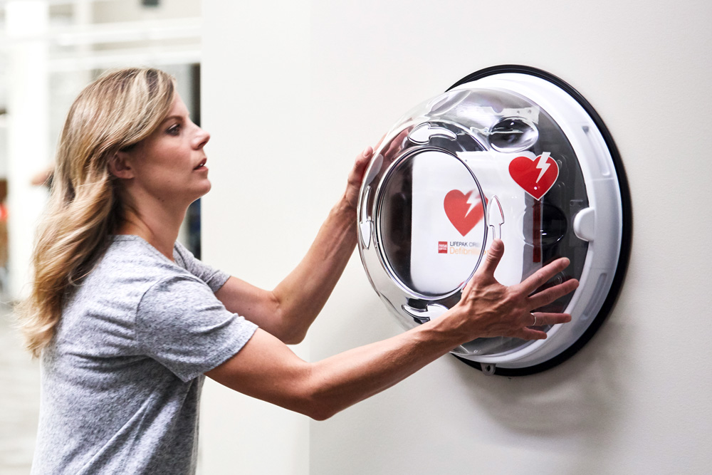En kvinna i grå t-shirt öppnar ett runt, väggmonterat skåp med genomskinligt lock som innehåller en hjärtstartare (defibrillator). Skåpet är märkt med röda hjärtsymboler med en blixt, vilket signalerar att det är en akut livräddningsutrustning. Miljön ser ut att vara en offentlig inomhusplats, som ett kontor eller en träningsanläggning.