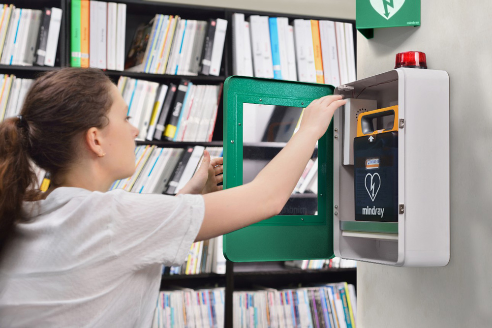 Kvinna öppnar ett väggskåp med en hjärtstartare av märket Mindray i en miljö som liknar ett kontor eller bibliotek, med bokhyllor i bakgrunden.