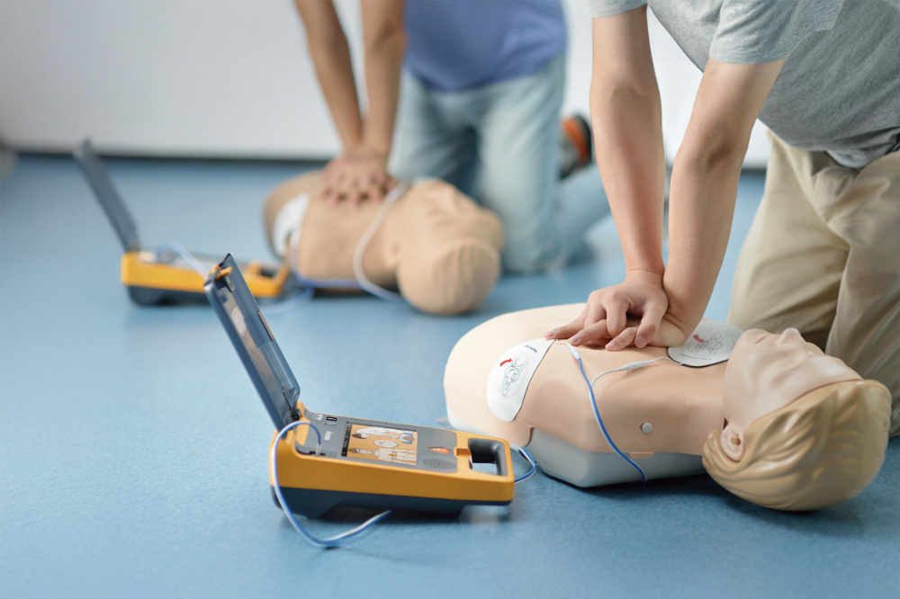 Två personer deltar i en HLR-kurs. I förgrunden utför en person bröstkompressioner på en HLR-docka bredvid en Mindray Benheart C2 defibrillator som är öppen och ansluten till dockan. I bakgrunden utför en annan person också bröstkompressioner på en liknande docka.