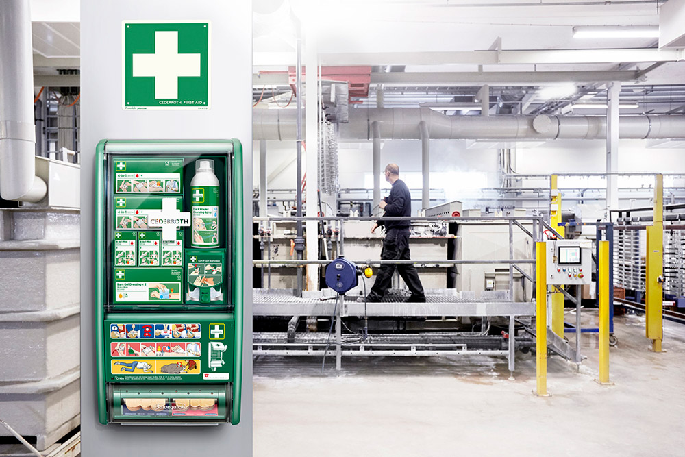 En första hjälpen-station från Cederroth är monterad på en vit pelare i en industrimiljö. Stationen innehåller olika förbandsmaterial, ögonskölj och instruktioner för akuta situationer. Ovanför finns en grön skylt med ett vitt kors som markerar platsen för första hjälpen. I bakgrunden syns en arbetare i mörka arbetskläder som går på en upphöjd metallplattform i en fabrikslokal med rör och maskinutrustning.