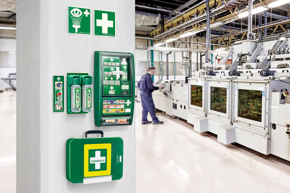 En pelare i en industrilokal är utrustad med flera första hjälpen-produkter från Cederroth, inklusive en ögonduschstation, en komplett första hjälpen-tavla samt en grön väska för akutvård. Ovanför finns skyltar med symboler för ögondusch och första hjälpen. I bakgrunden arbetar en person i blå arbetskläder vid en stor industrimaskin i en ljus och modern fabriksmiljö.