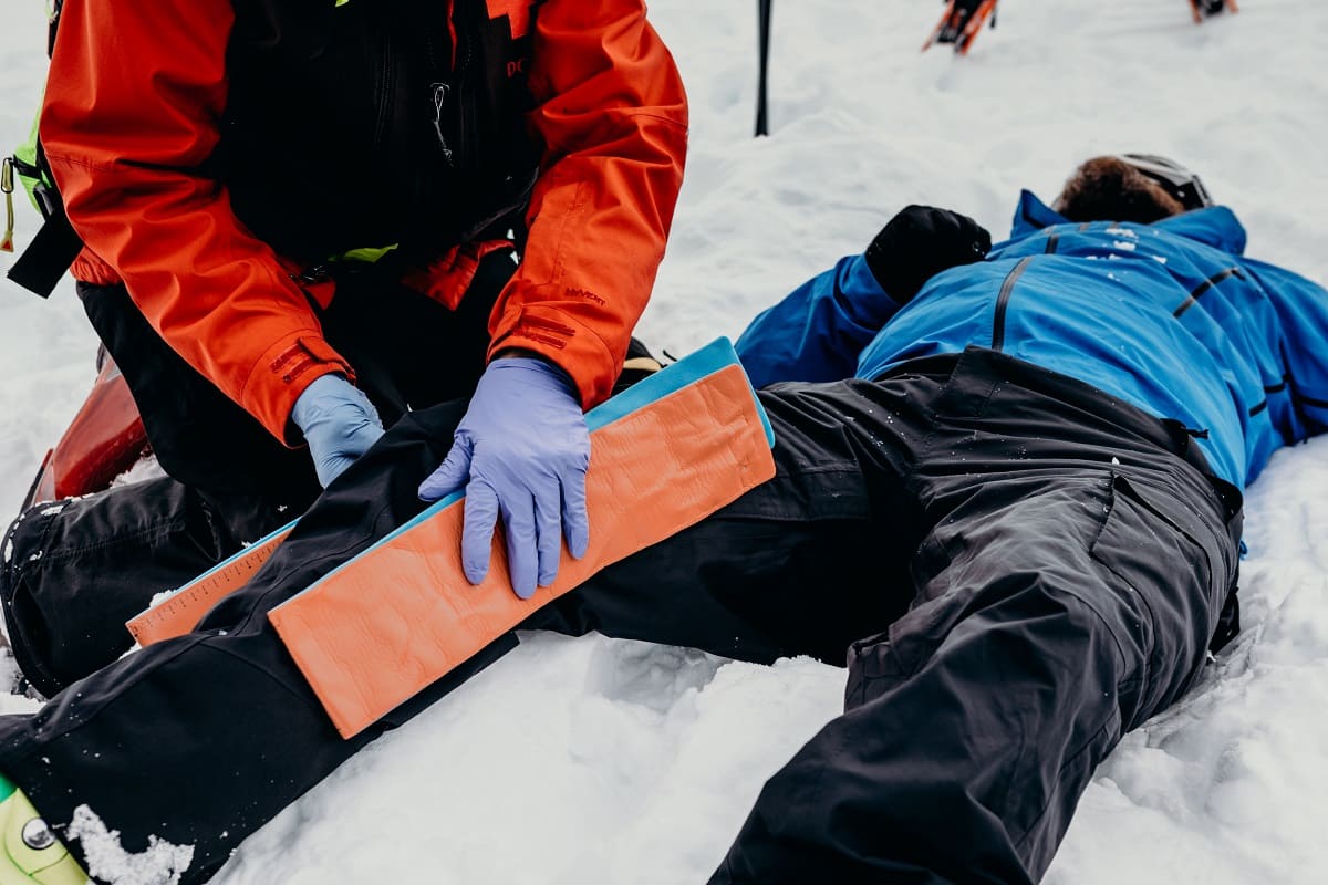 En person i röd jacka och blå handskar knäböjer i snön och håller en orange och blå splint på benet av en person i blå jacka som ligger ner.