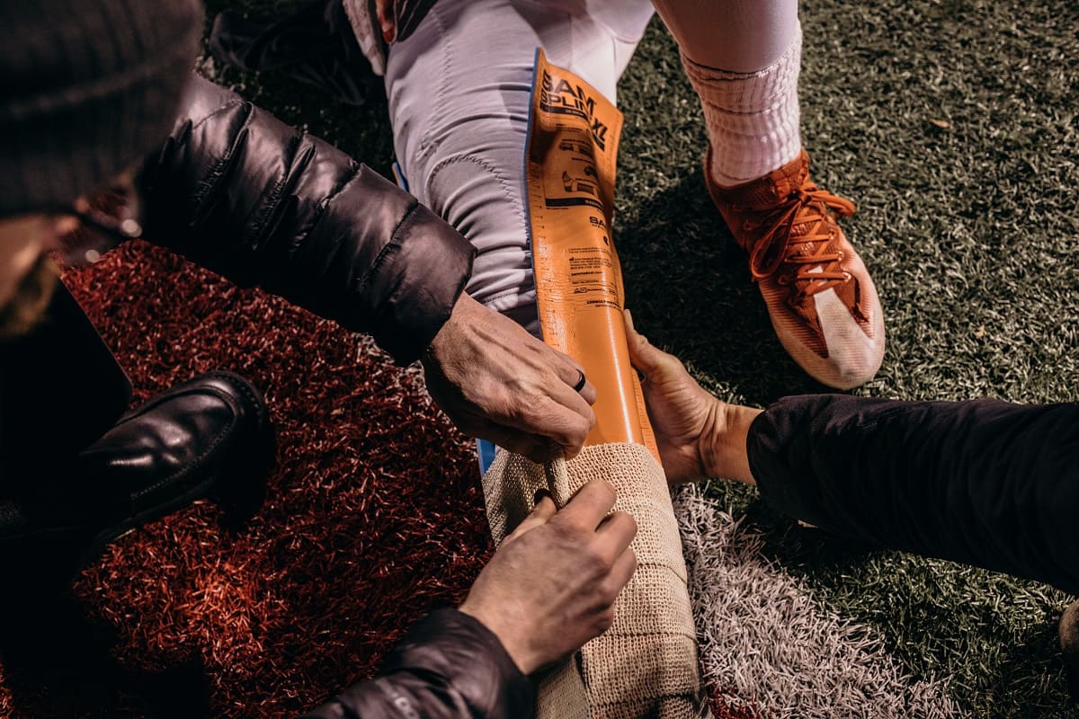 En närbild av händer som håller en orange SAM Splint på benet av en amerikansk fotbollsspelare, medans ett bandage och trauma förband lindas runt.