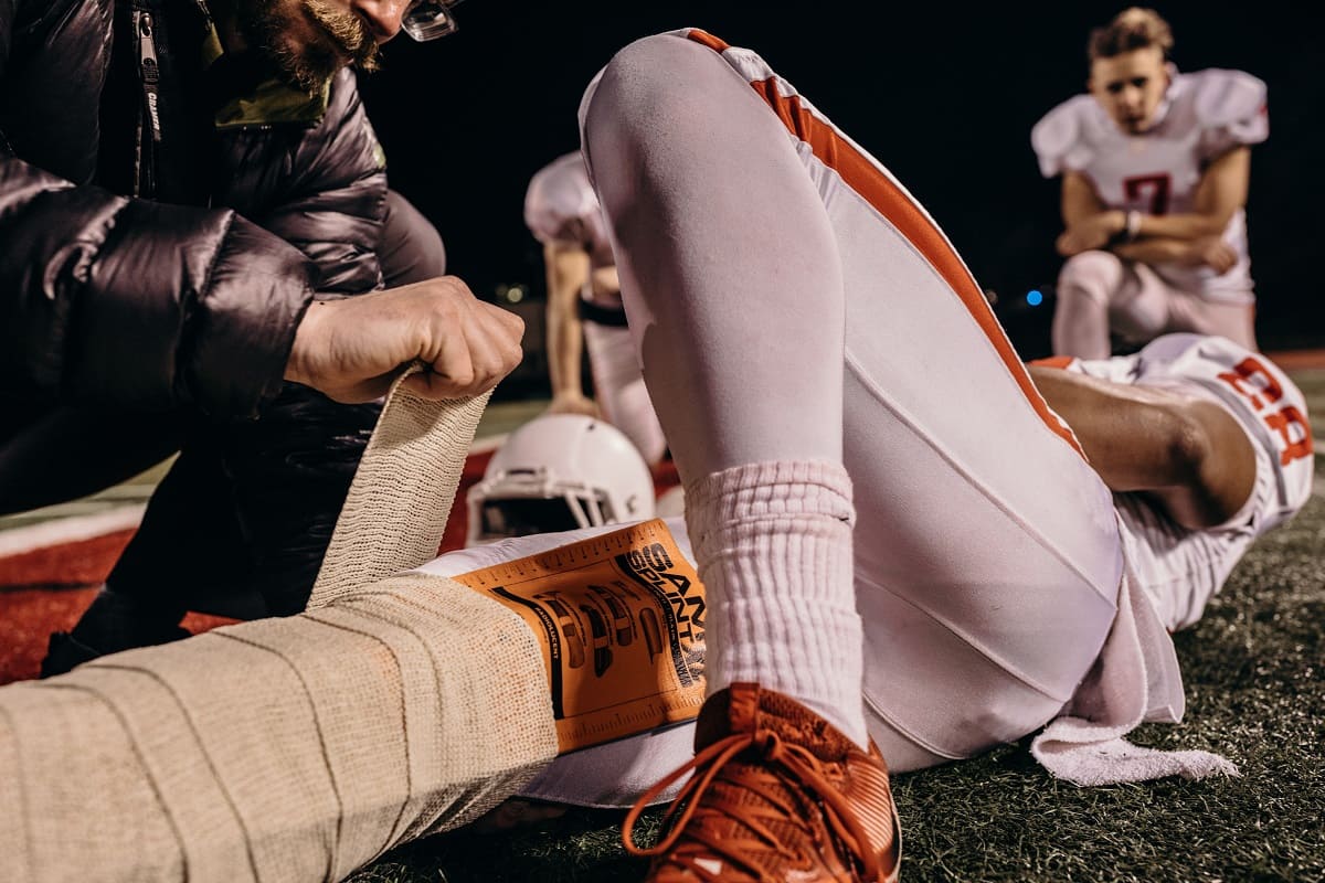 Flera personer assisterar en amerikansk fotbollsspelare som ligger på marken som får en SAM Splint applicerad på benet med ett trauma förband. Ytterligare en spelare sitter i bakgrunden.