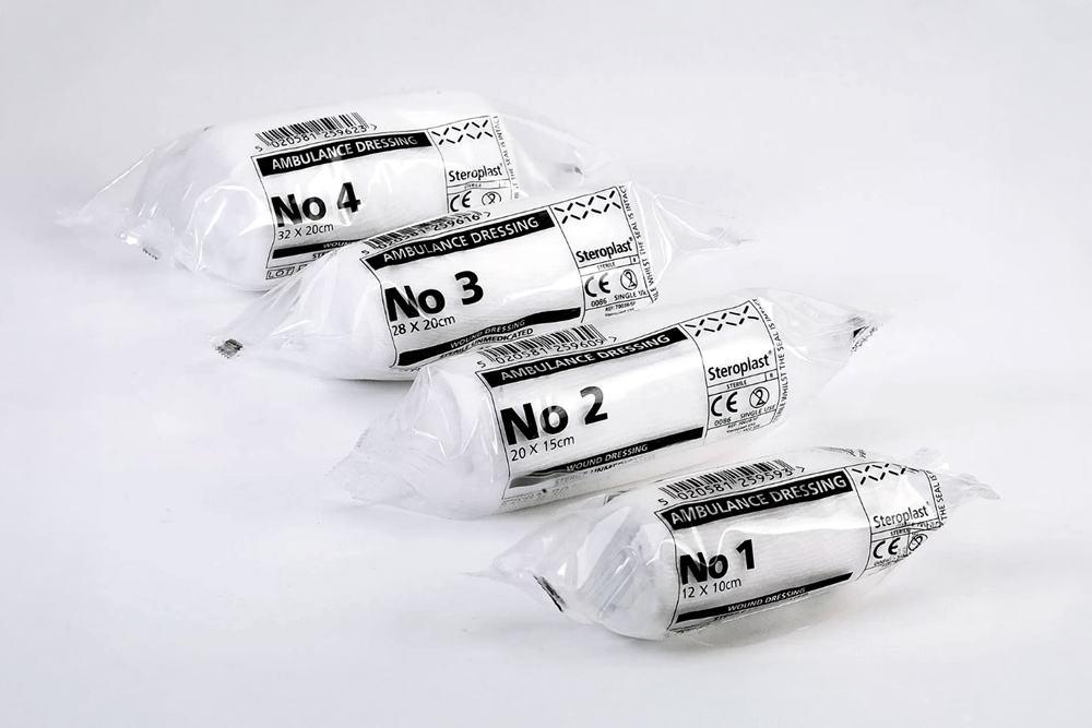 Fyra individuellt förpackade vita "Ambulance Dressing" förband i olika storlekar (No 1, No 2, No 3, No 4), arrangerade diagonalt på en vit bakgrund.