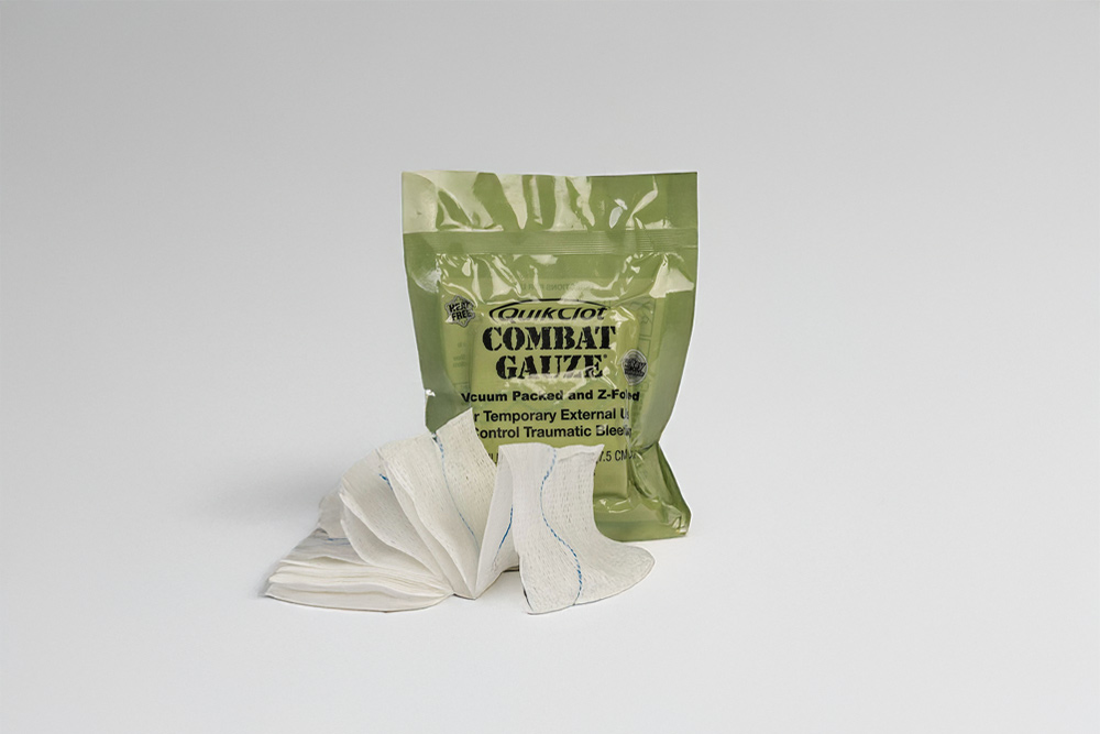 En senaps grön förpackning av "QuikClot Combat Gauze" med en del av gasväven utfälld framför den, på en vit bakgrund.